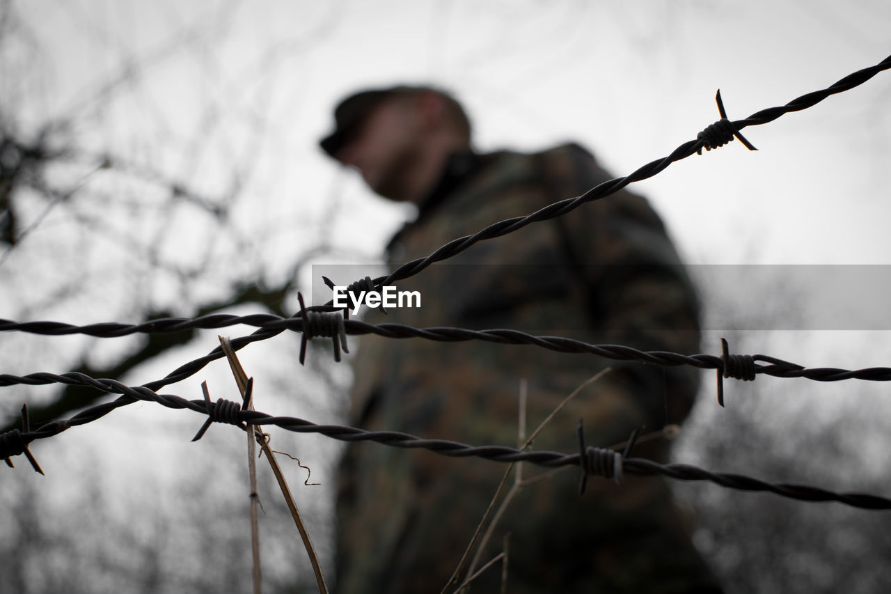 Close-up of barbed wire fence with man standing in background against sky