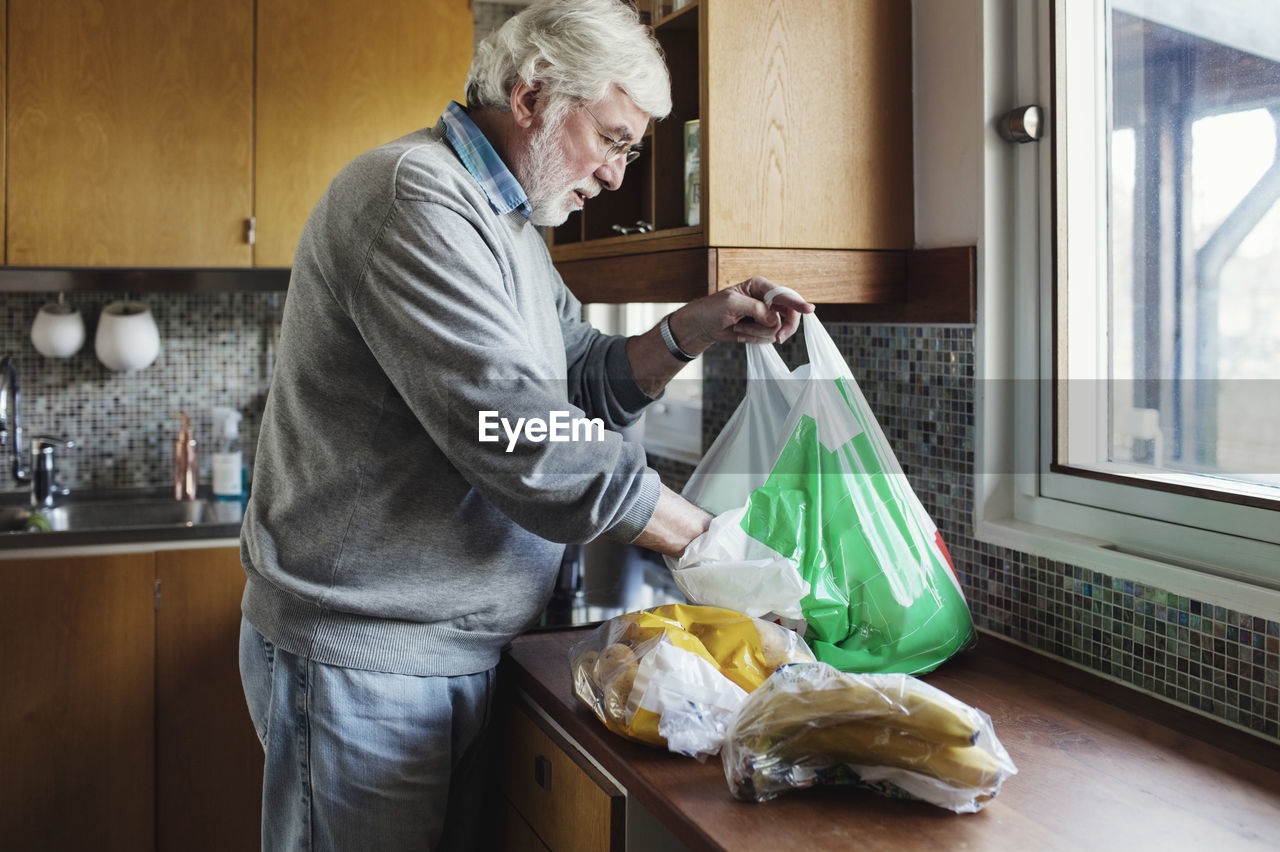Senior man removing packets from plastic bag at kitchen counter