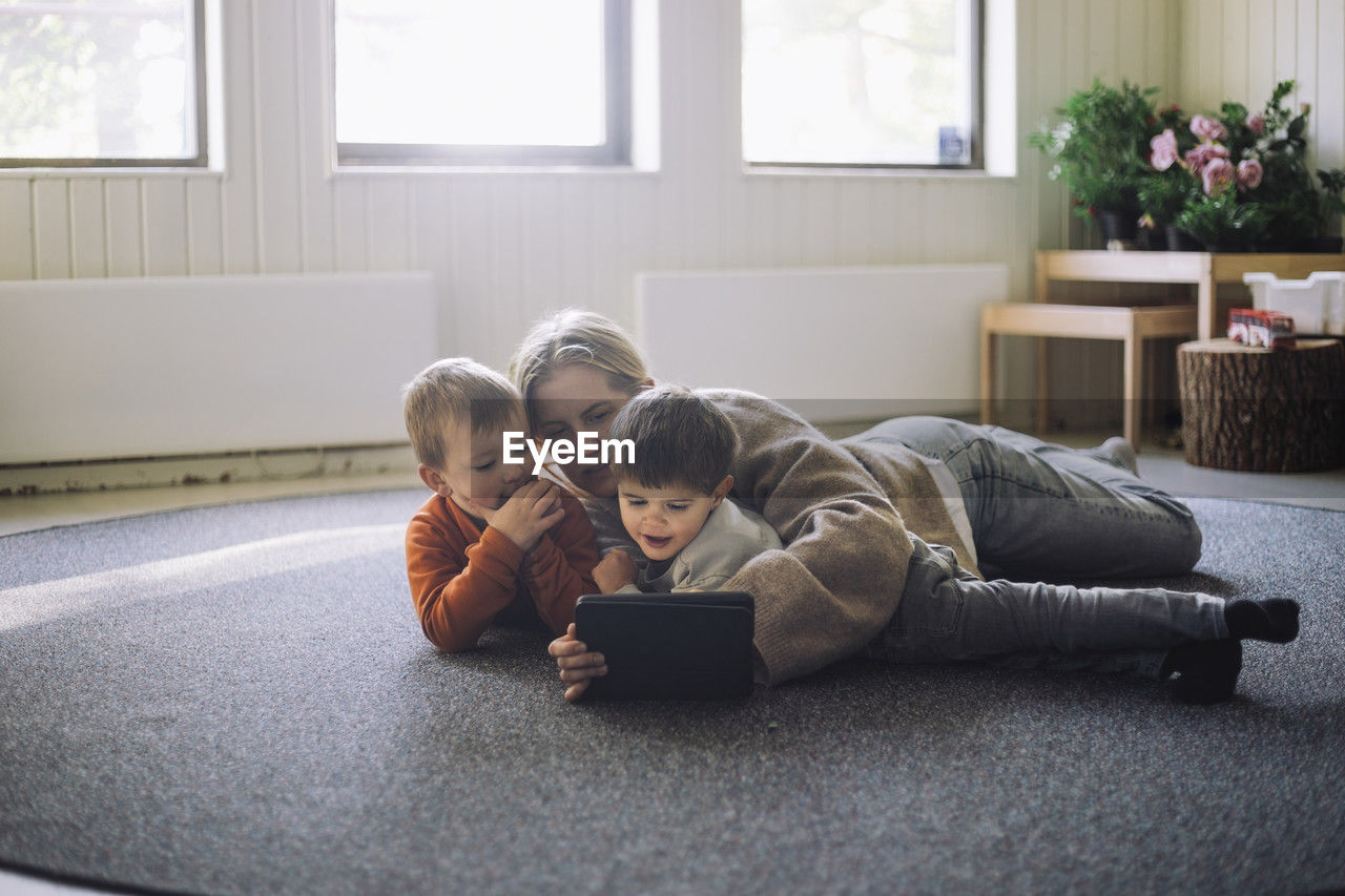 Female teacher watching digital tablet with boys while lying down on carpet at preschool