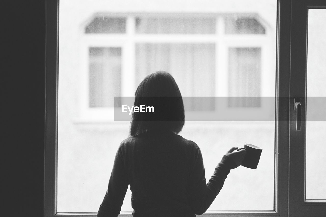 Rear view of woman holding cup by window