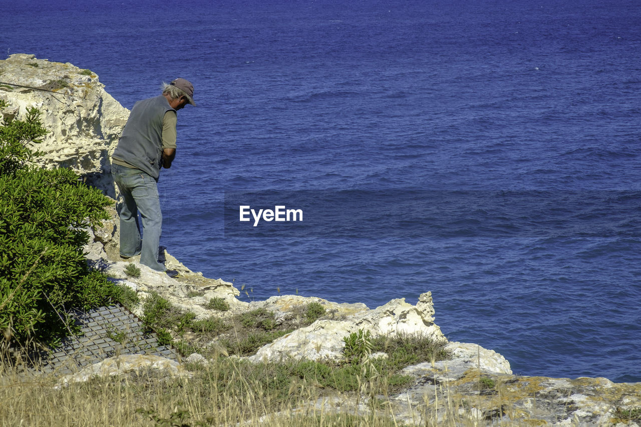 MAN STANDING ON ROCK BY SEA AGAINST SKY