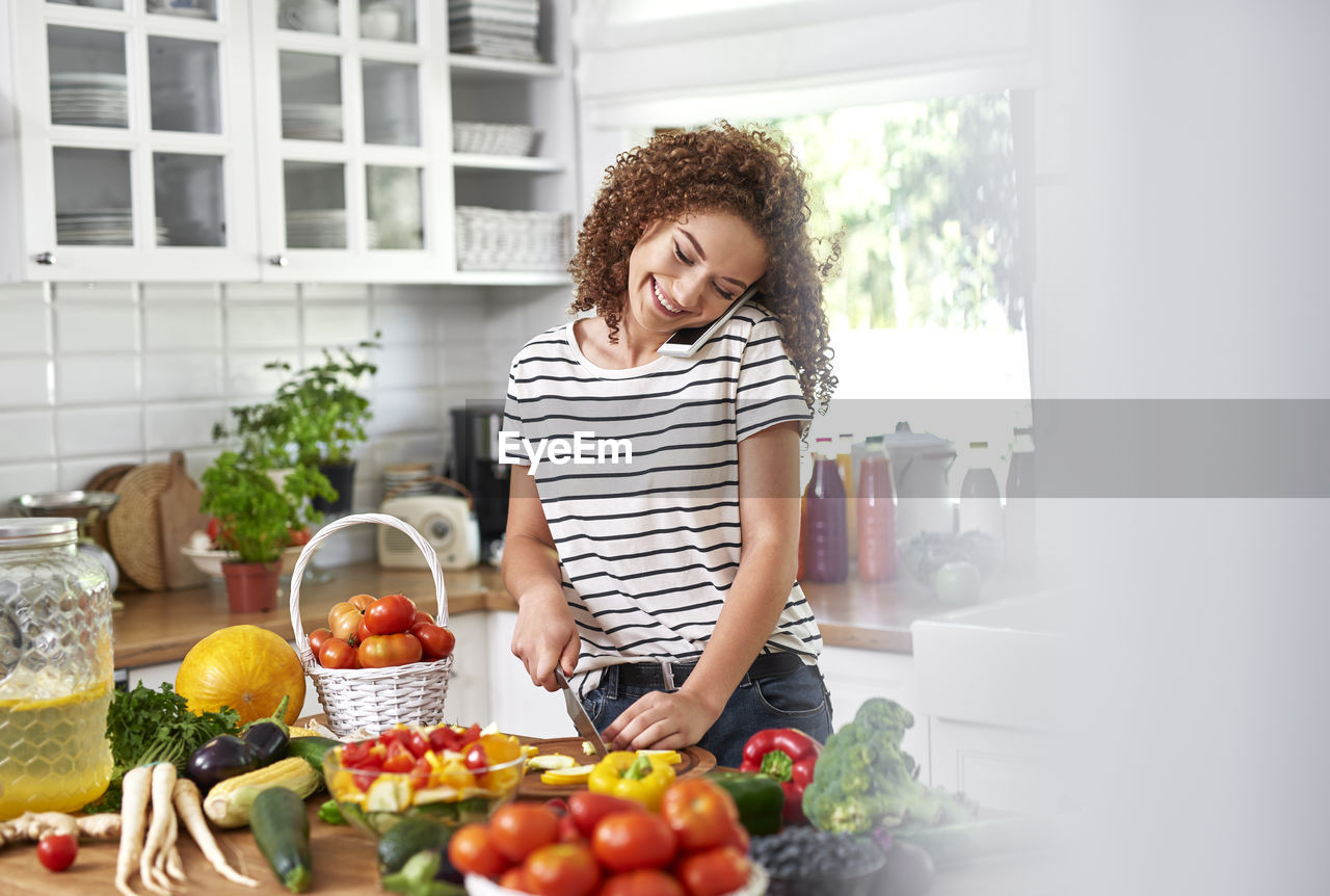 Smiling teenage girl talking over smart phone while standing in kitchen at home