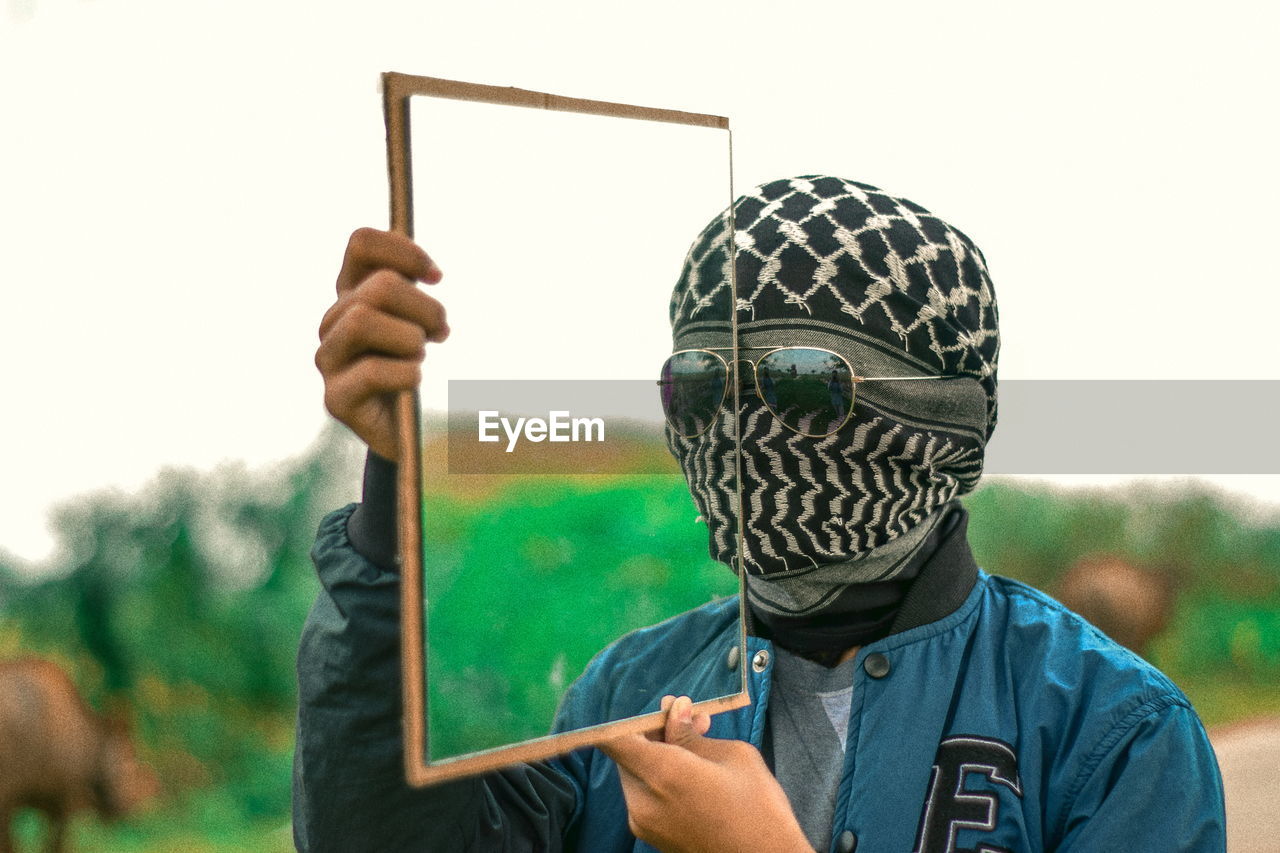 Portrait of young man with covered face holding mirror against clear sky