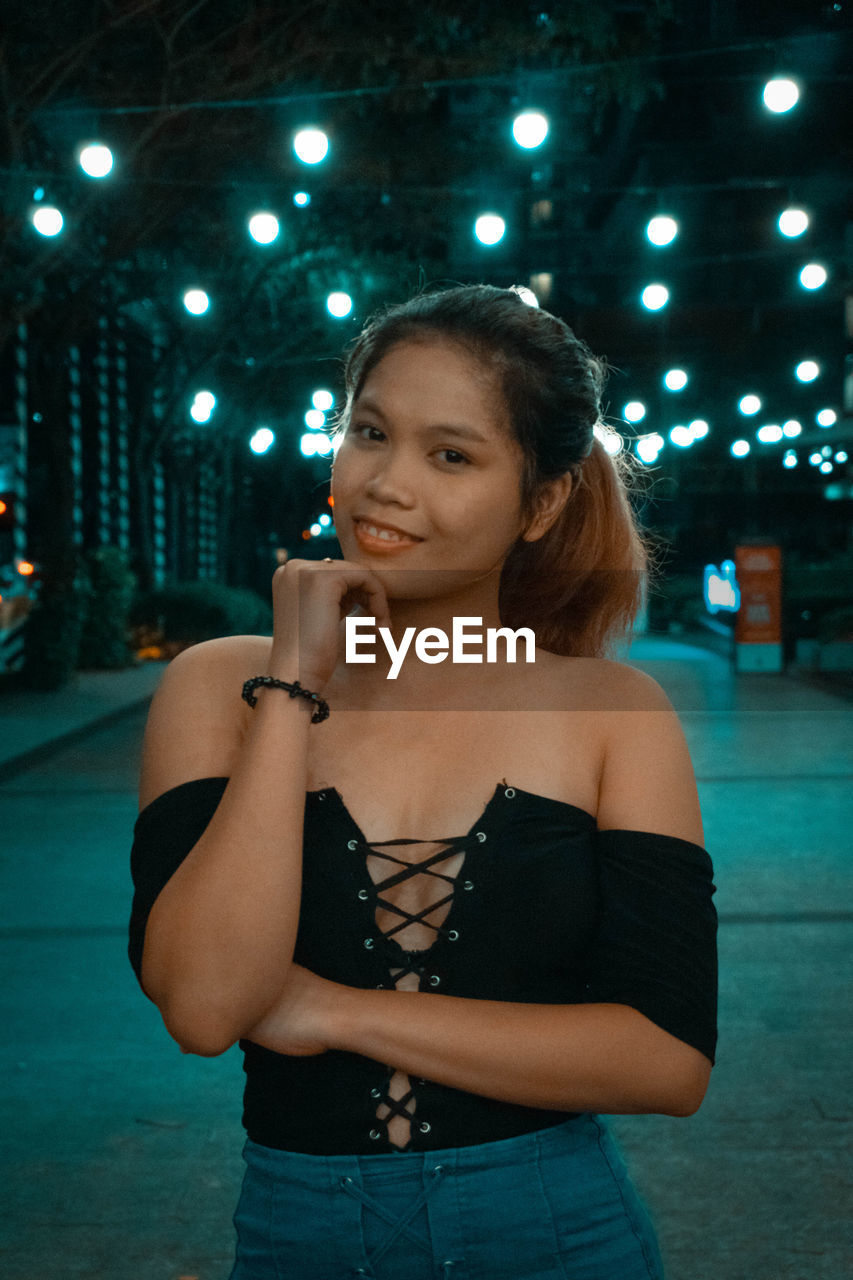 PORTRAIT OF YOUNG WOMAN STANDING AGAINST ILLUMINATED LIGHTS