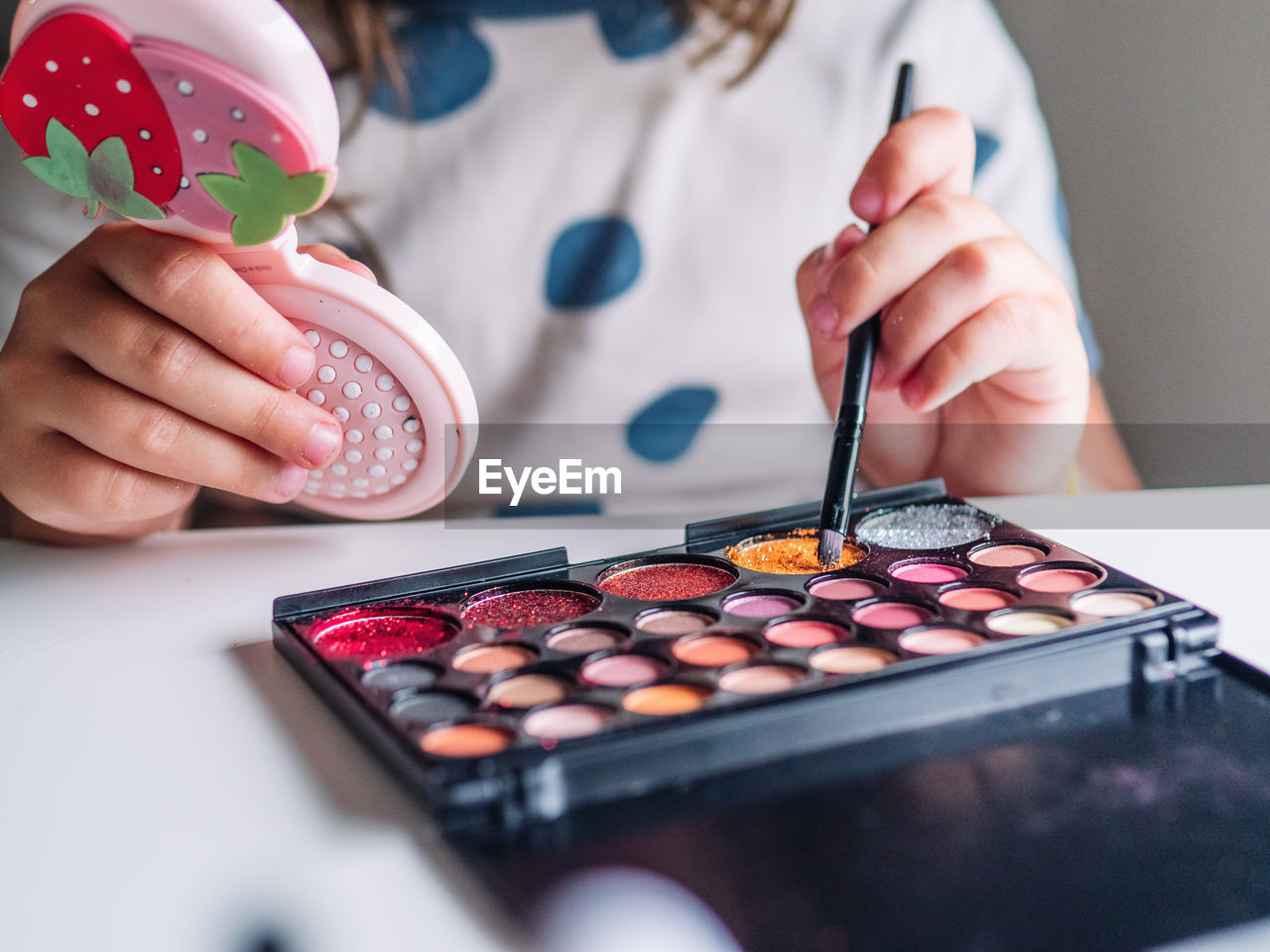 Crop unrecognizable kid with applicator and mirror making up face at table with assorted cosmetic products in house