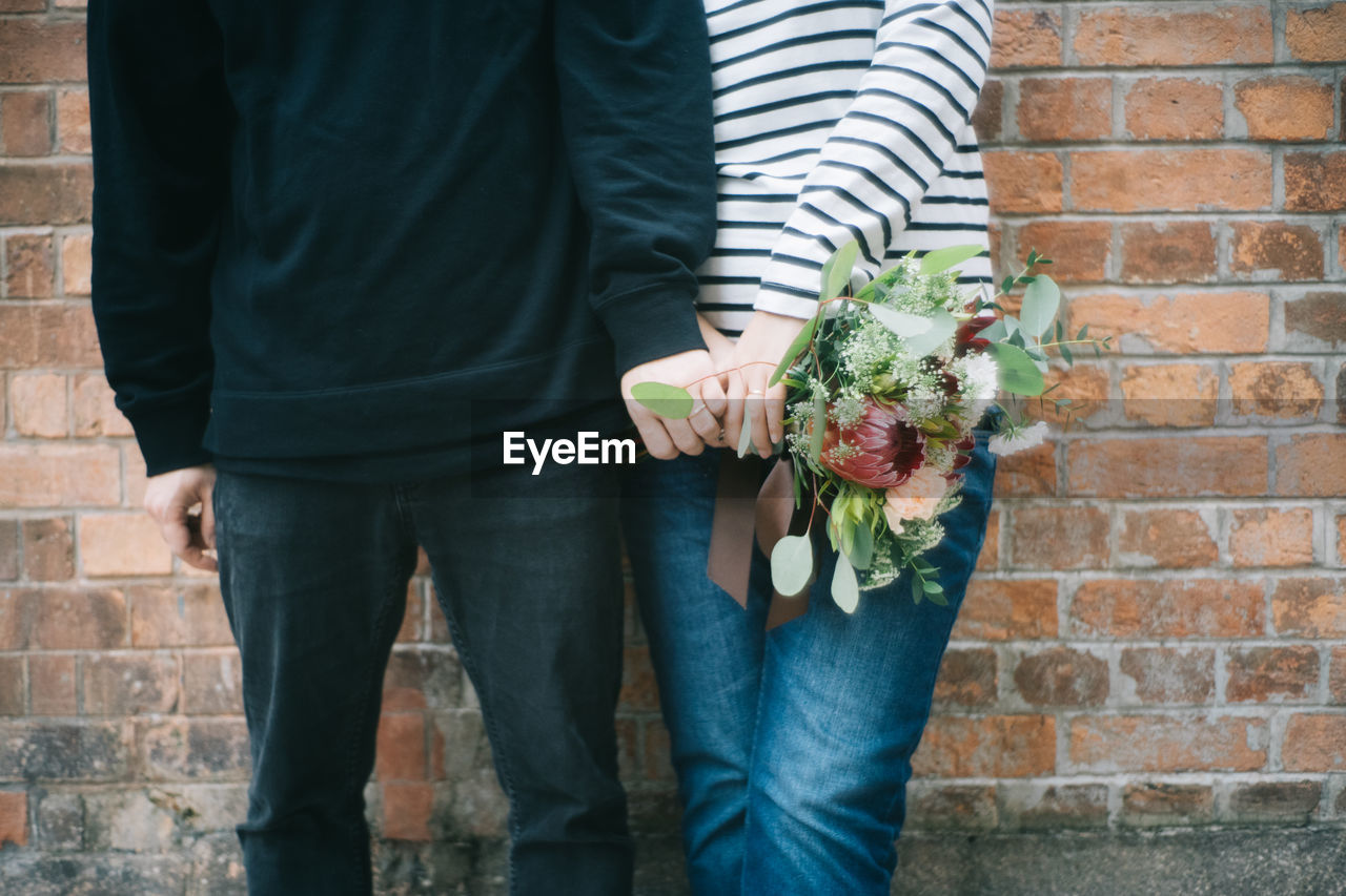 Midsection of couple with bouquet standing against brick wall