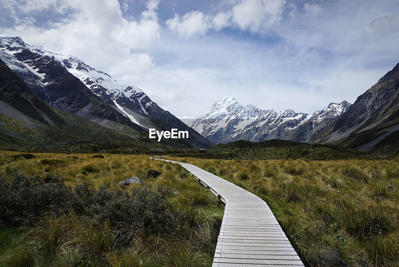 Boardwalk leading towards snowcapped mountains against cloudy sky