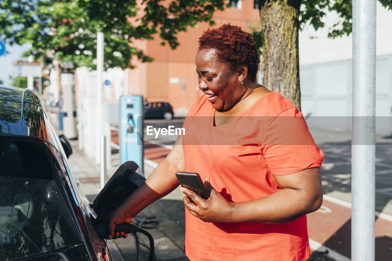 Happy woman holding mobile phone charging electric car on sunny day