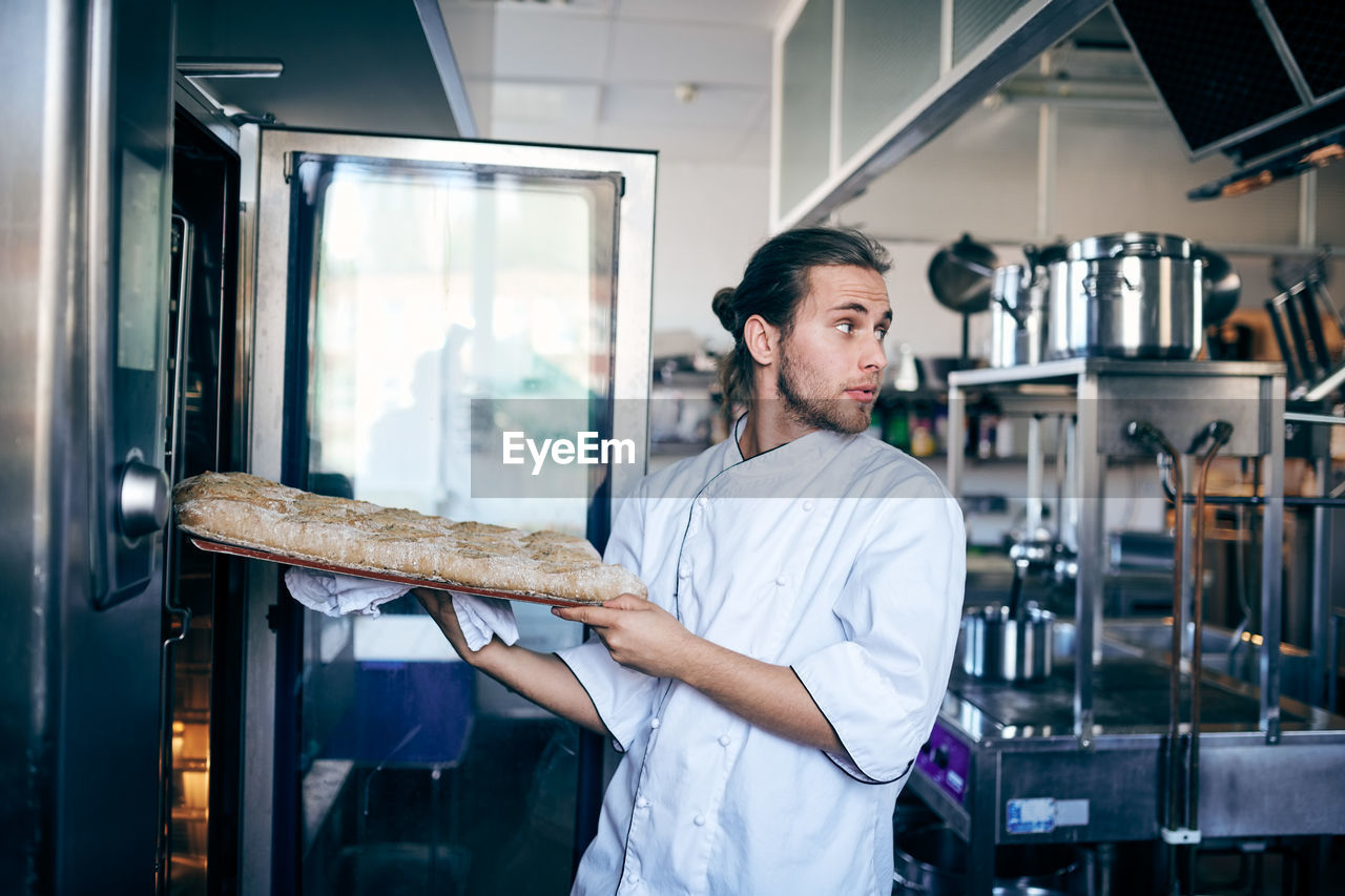 Male chef putting bread in oven while looking away at commercial kitchen