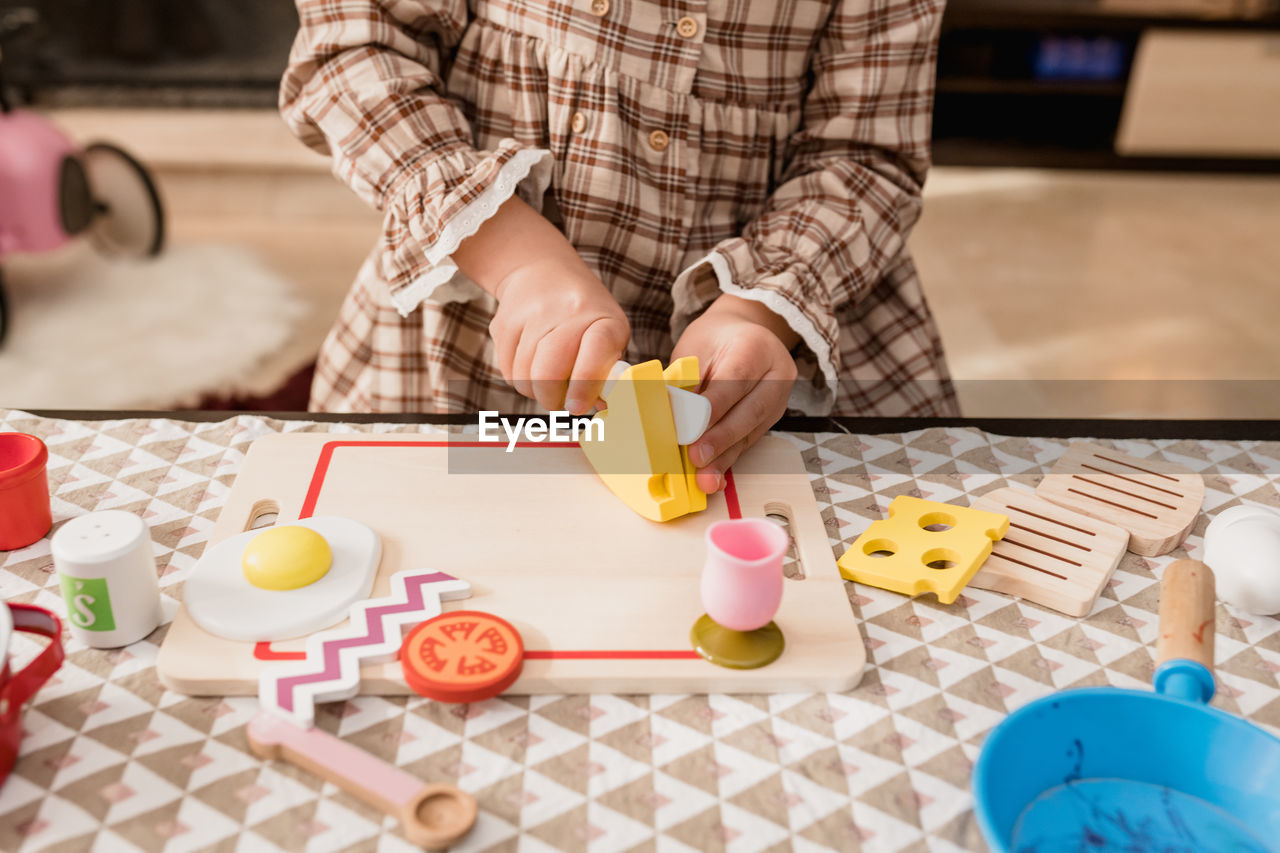 Crop unrecognizable kid in checkered dress with toy knife cutting cheese on chopping board while playing in house