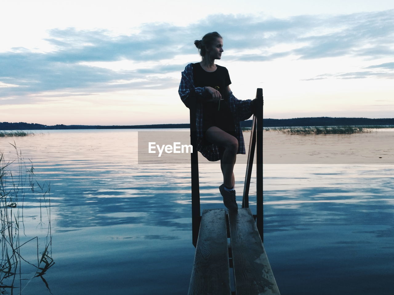 Young woman standing on wooden plank in lake against sky during sunset