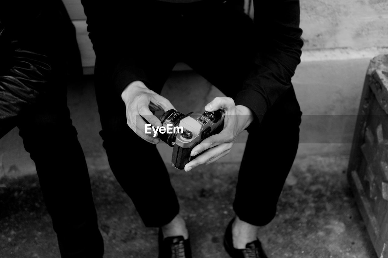 Low section of man holding camera while sitting on retaining wall