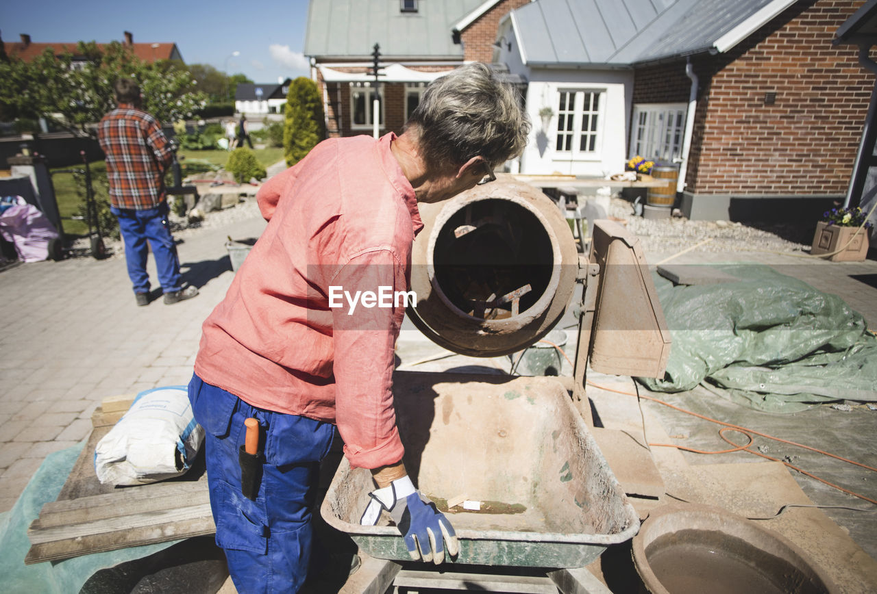 Side view of senior woman using cement mixer while man standing in background at yard