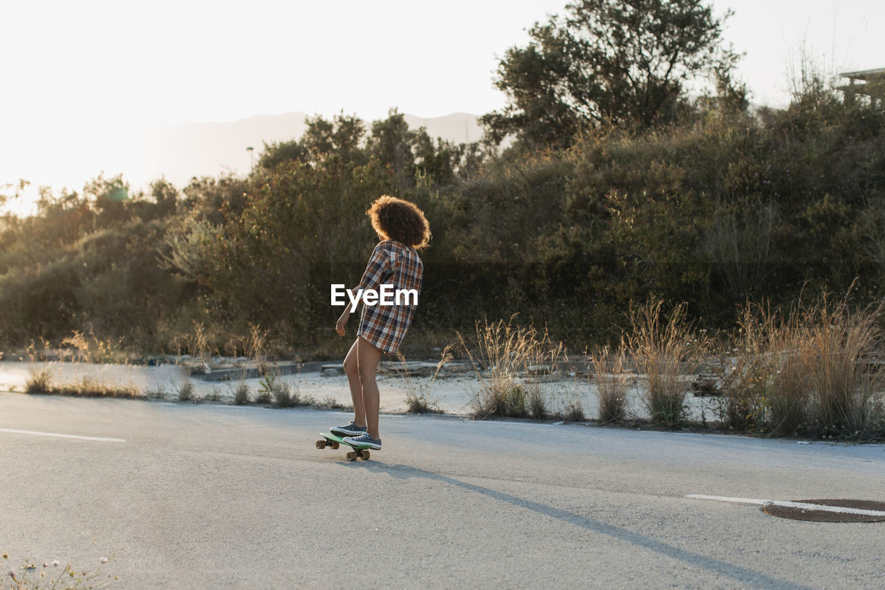 Back view of female hipster with afro hairstyle riding penny board along empty road during sunset