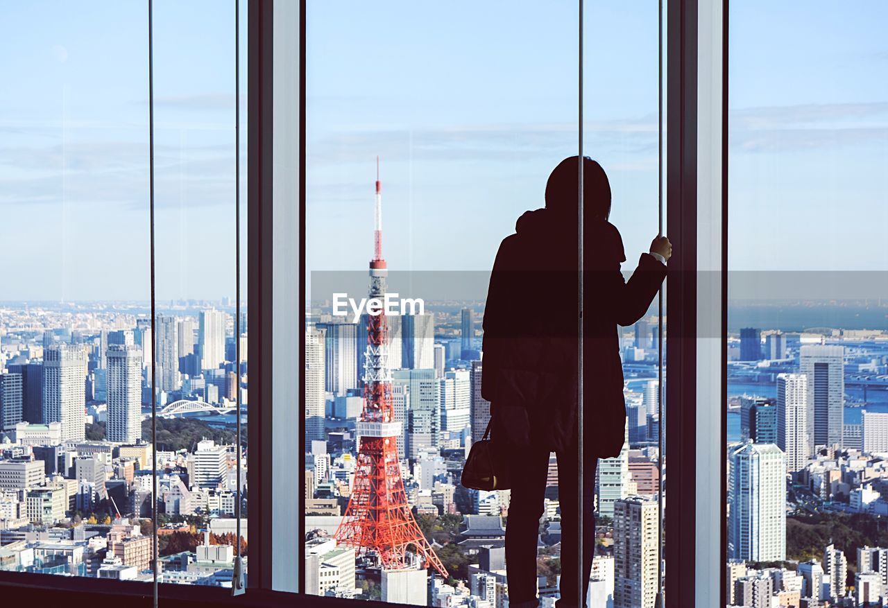 Rear view of woman standing by window against tokyo tower in city