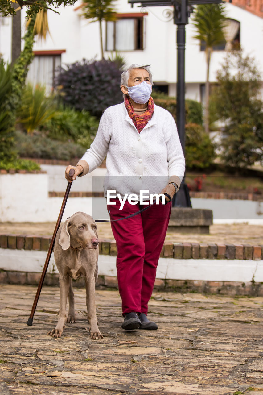 Full length of senior wearing mask with dog walking outdoors