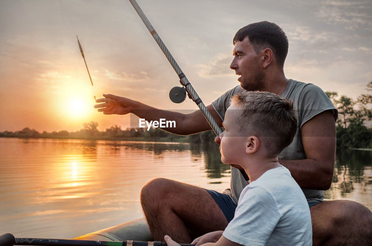 Happy father and son together fishing from a boat at sunset time, summer day, beautiful sky on lake.
