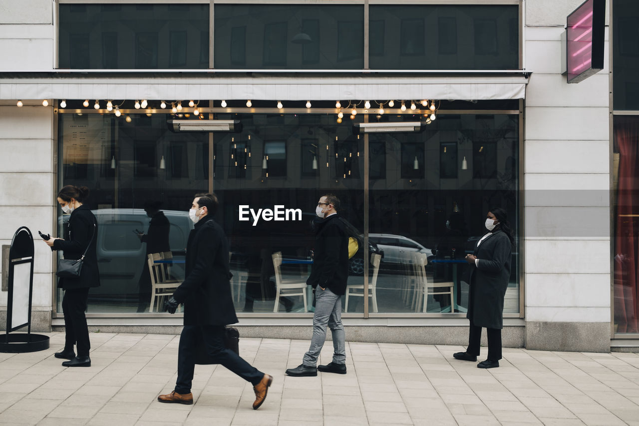 Businessman walking while male and female colleagues standing in line outside store during pandemic