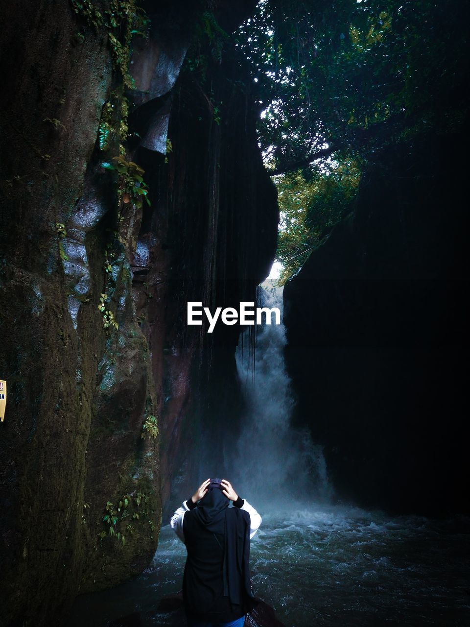 REAR VIEW OF MAN STANDING ON ROCK FORMATION IN WATER