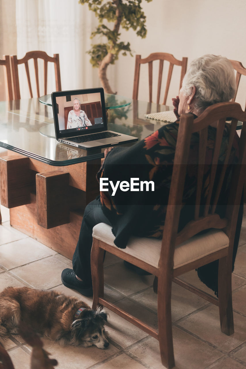 Back view of unrecognizable elderly female with short gray hair sitting on chair and listening goodbye of sister while making video call via netbook at home