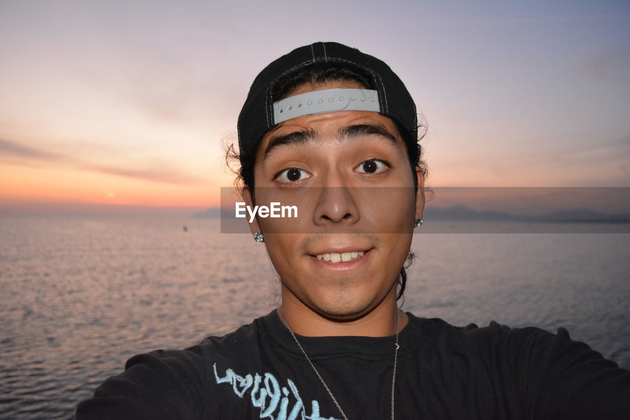 Close-up portrait of man wearing cap against sea during sunset