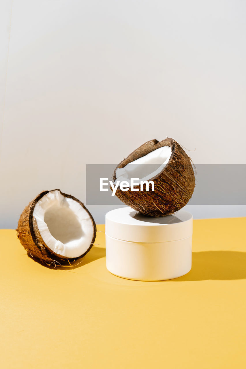 Two halves of coconut and a jar of cosmetics on a yellow and gray background.