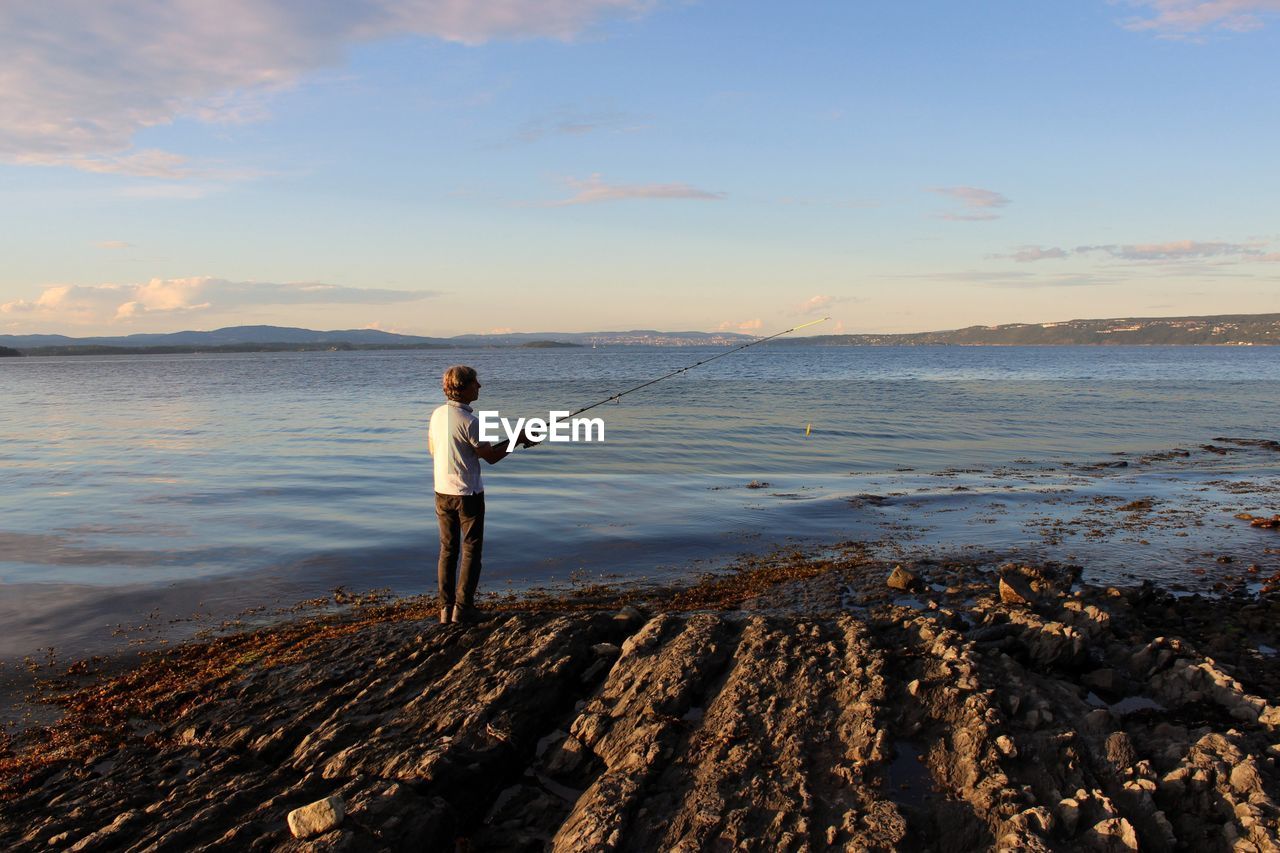 Rear view of man fishing while standing on shore at beach against sky