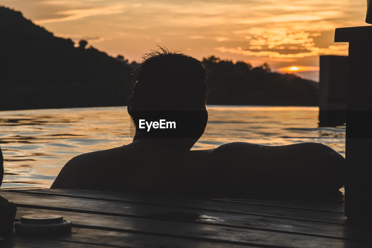 PORTRAIT OF SILHOUETTE MAN SITTING BY LAKE AGAINST ORANGE SKY