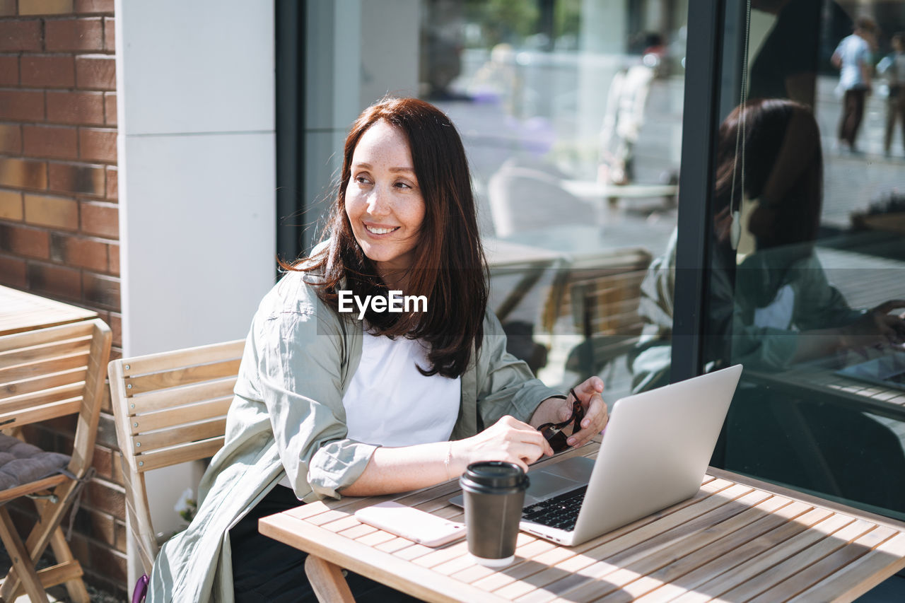 Adult smiling brunette business woman forty years in stylish shirt working on laptop in cafe 