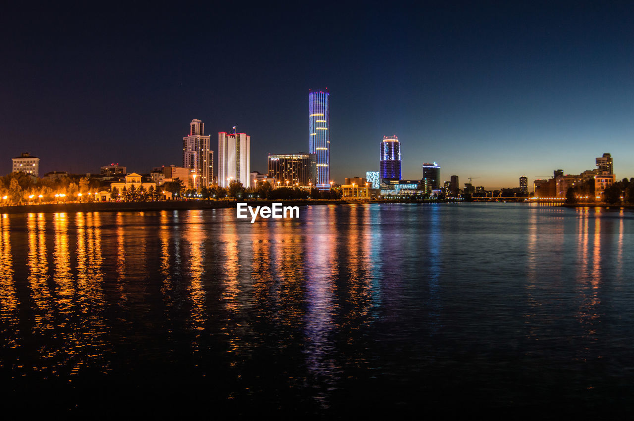 Illuminated buildings by river against clear sky at night