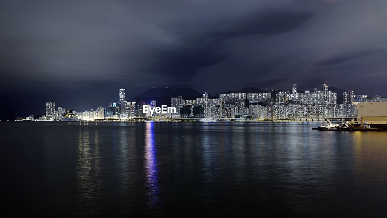 Illuminated cityscape in front of sea against cloudy sky at night