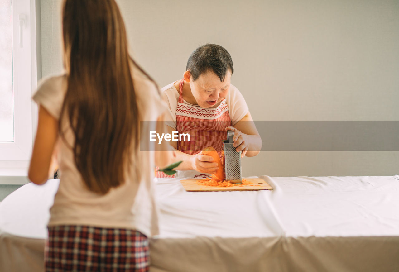 Lifestyle, education. an elderly woman with down syndrome rubs carrots on a grater with an assistant