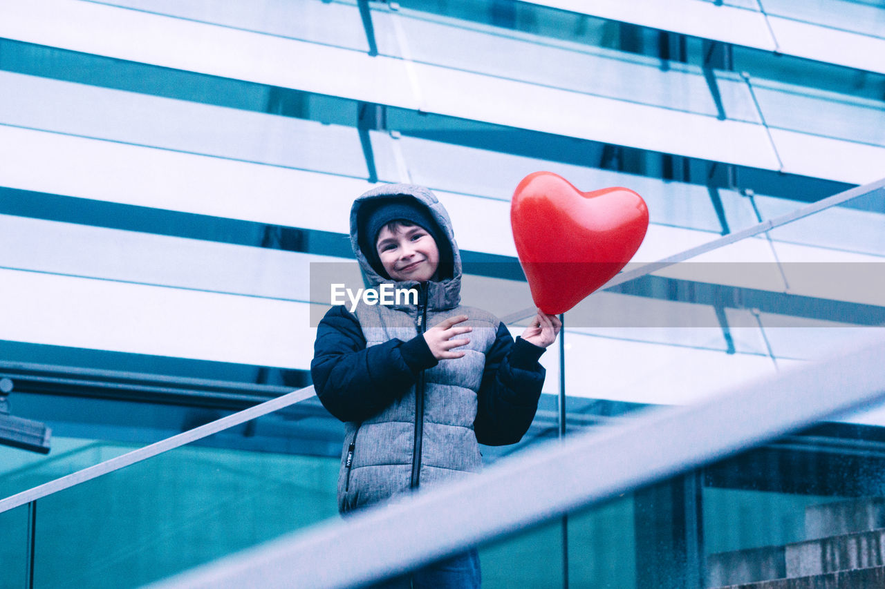 Low angle portrait of girl holding red balloon while standing against building
