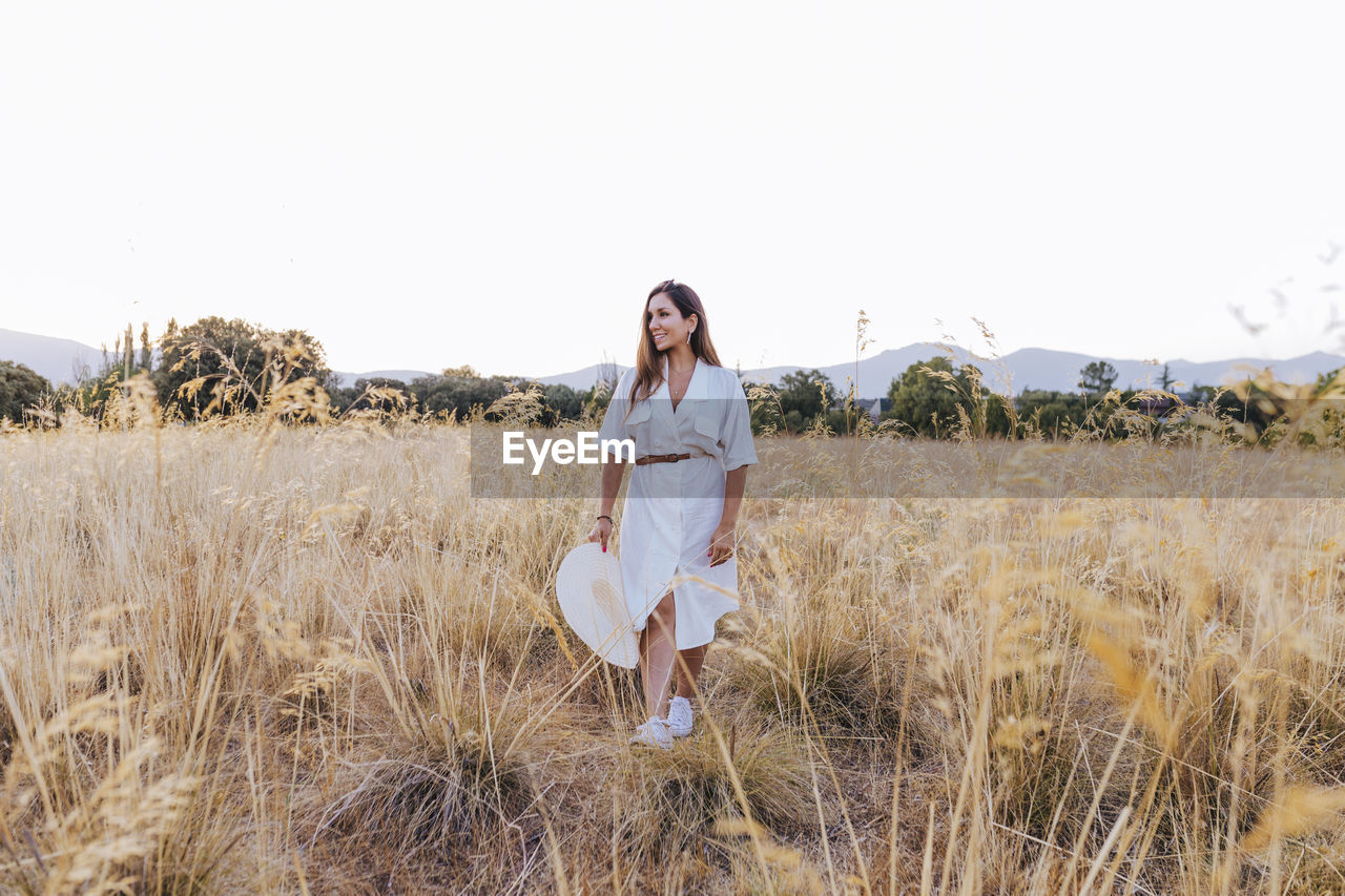 Smiling woman holding hat while standing on land against clear sky