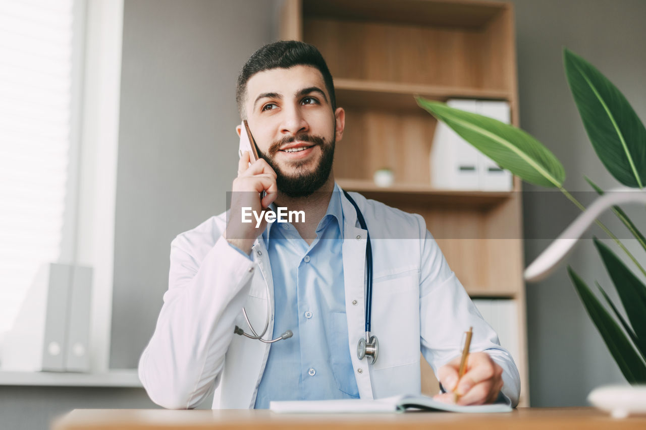 A young doctor of oriental appearance is talking on the phone, sitting at the table 