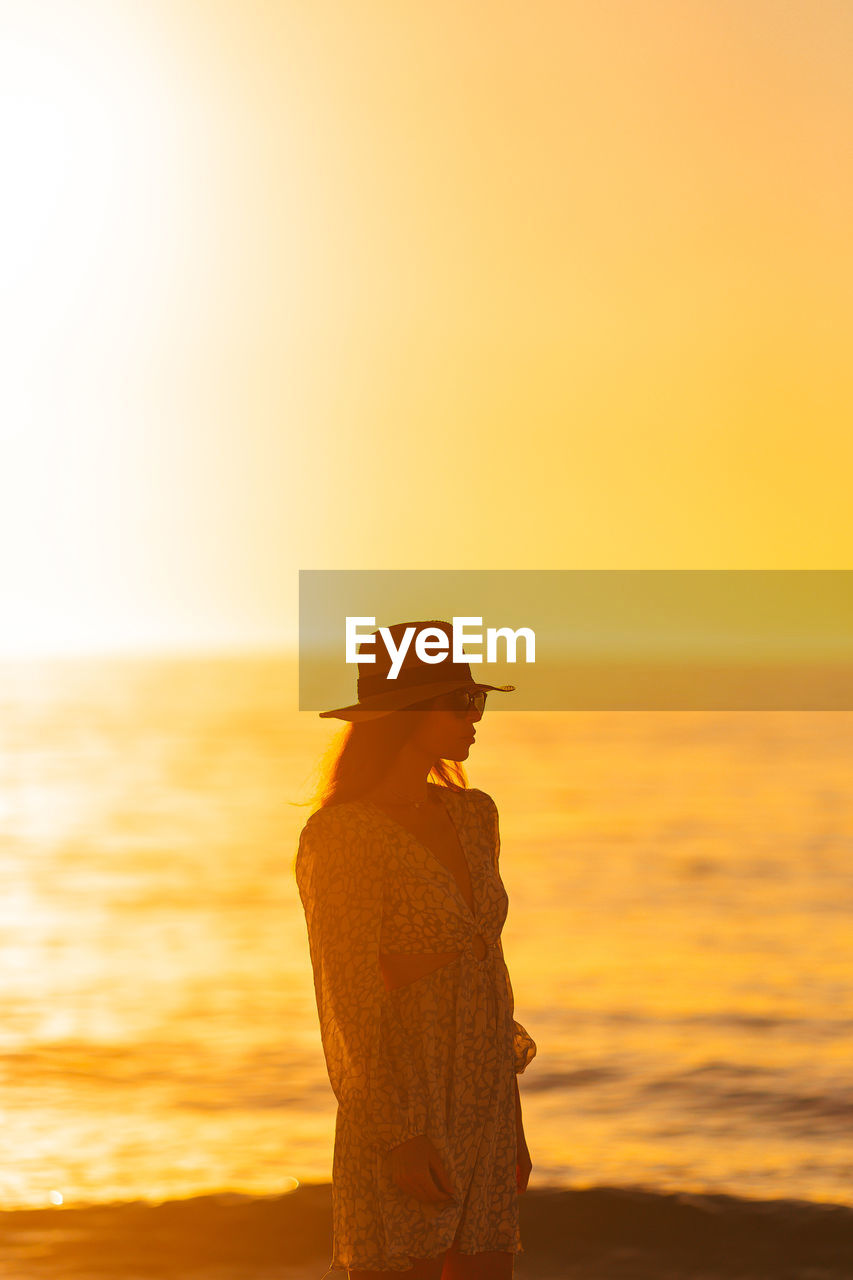 rear view of woman standing at beach against sky during sunset