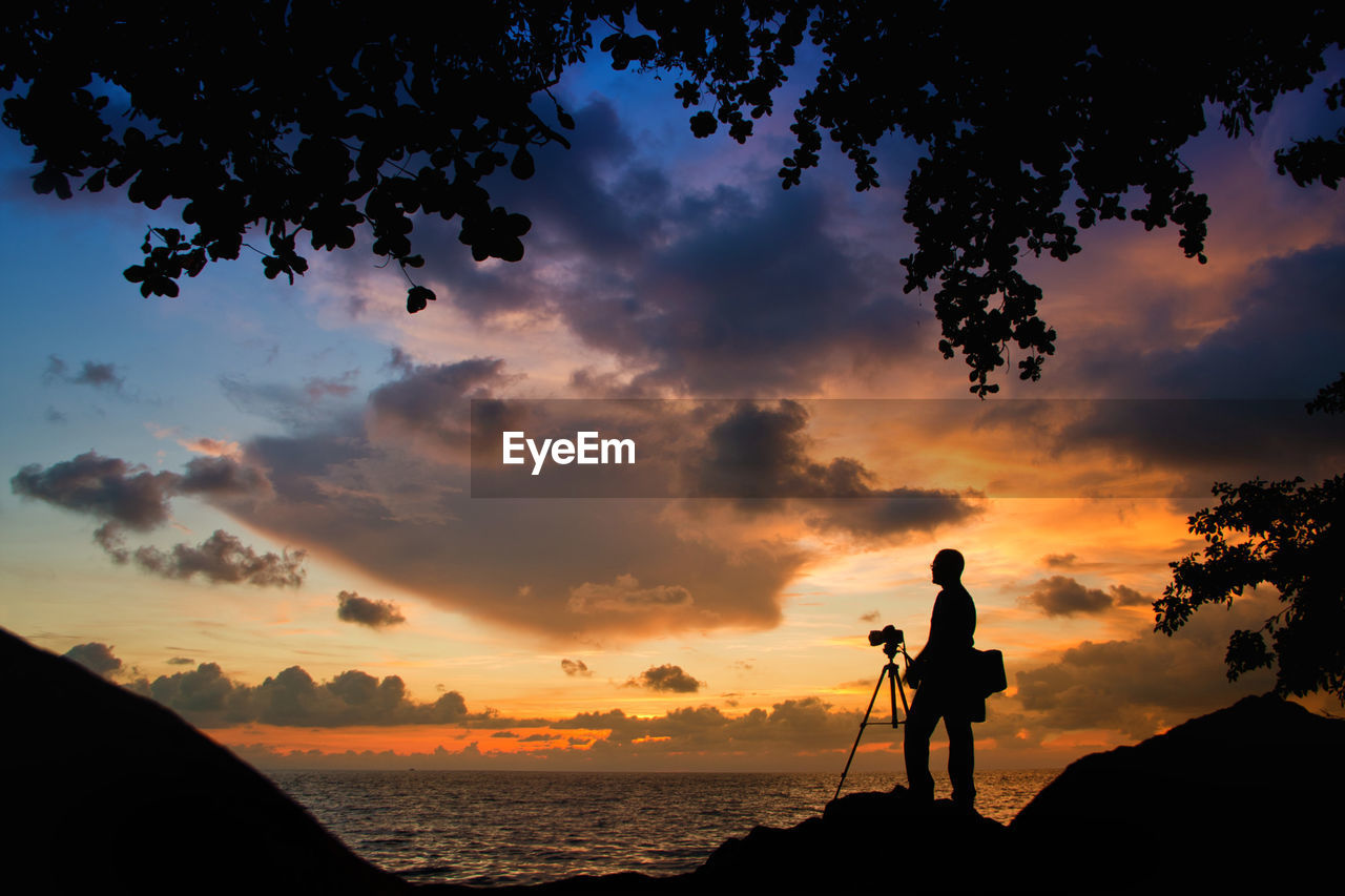 Silhouette man with camera on rock by sea against sky during sunset