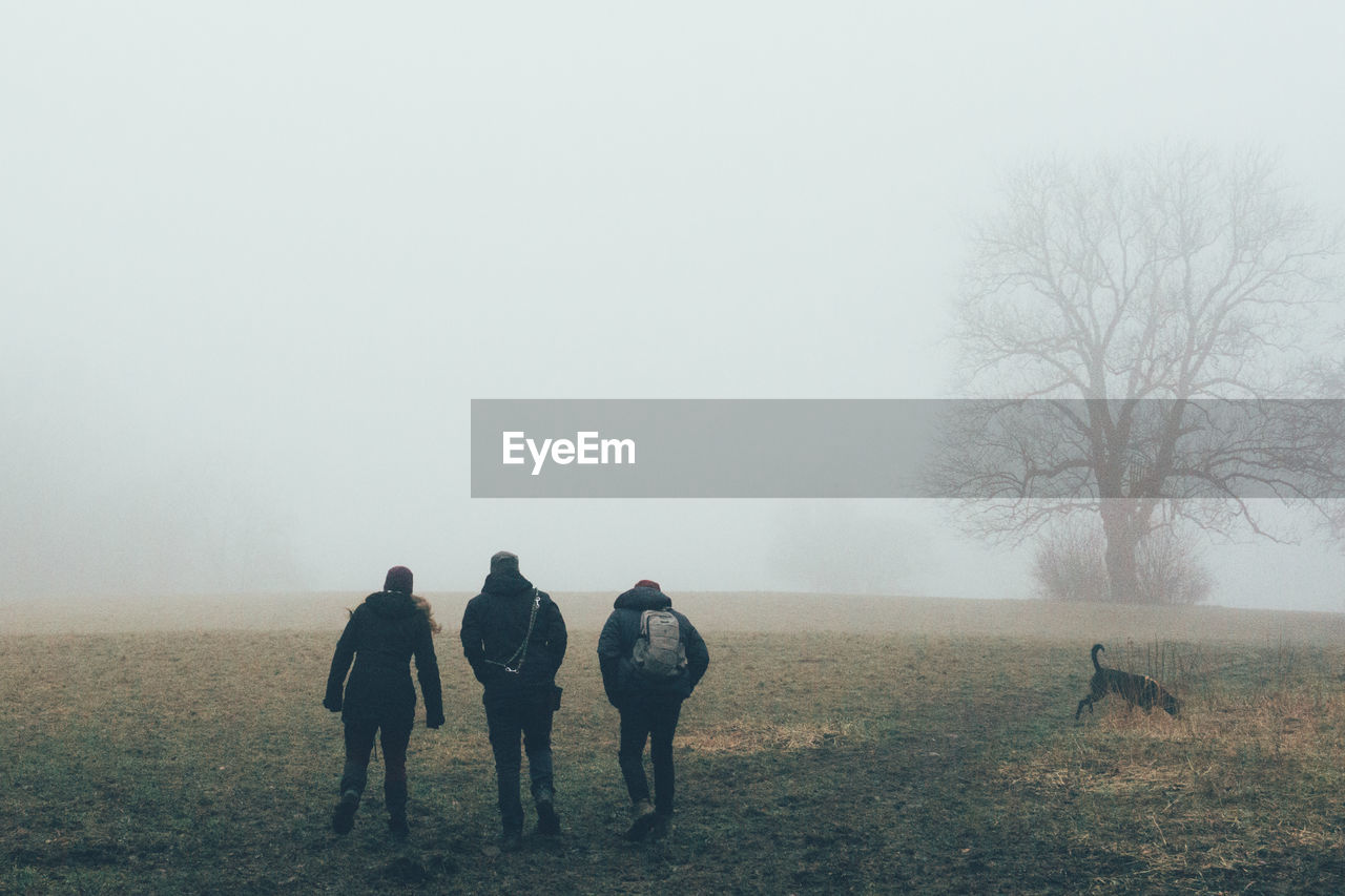 REAR VIEW OF PEOPLE WALKING ON FIELD AGAINST SKY DURING FOGGY WEATHER