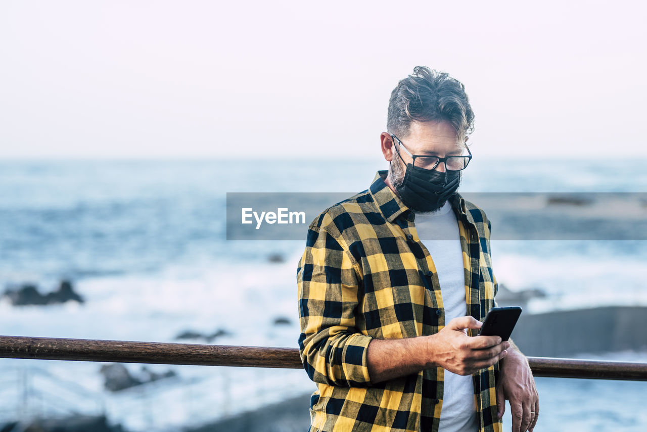 Mature man wearing mask using smart phone while standing against sea