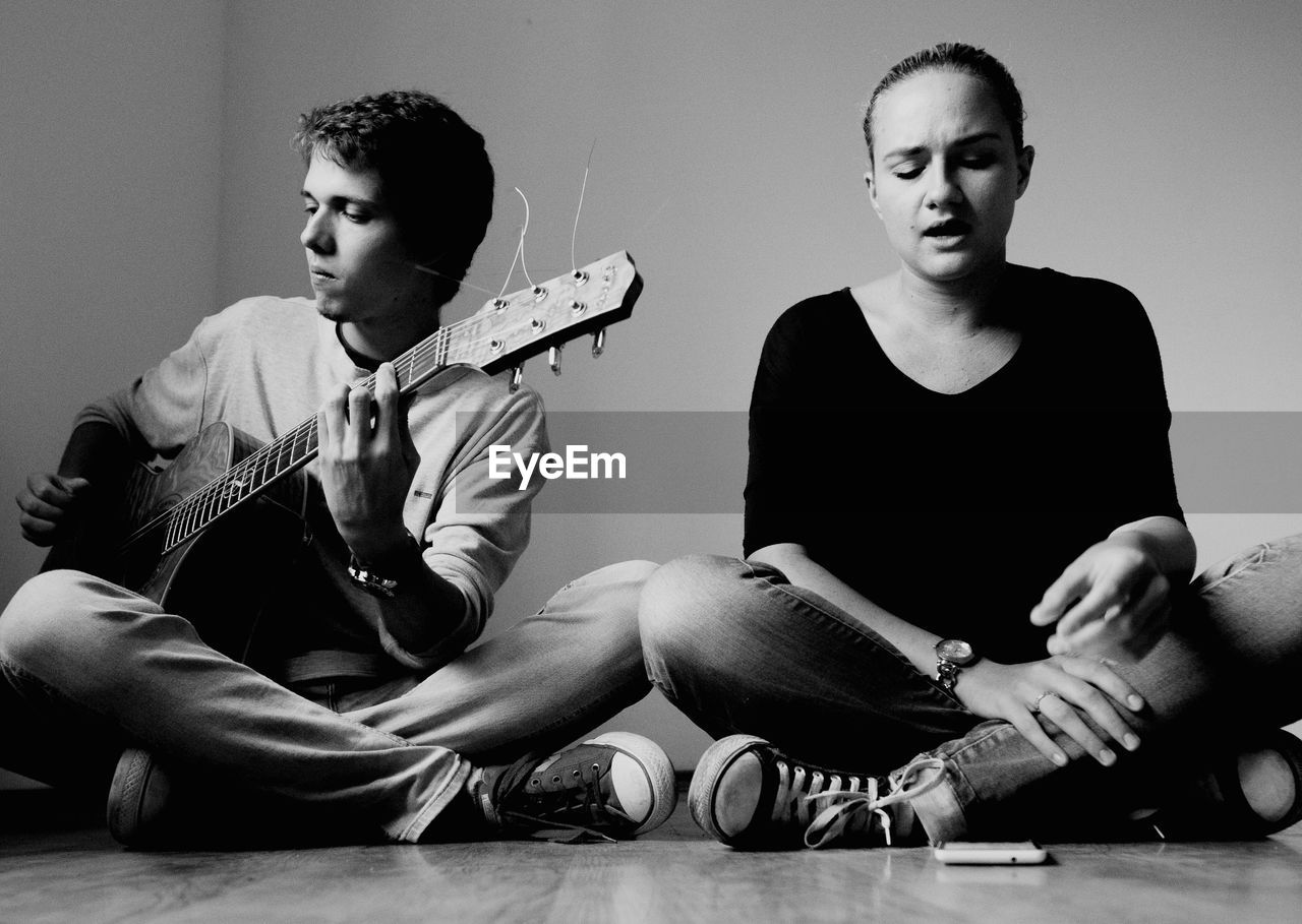 Young man with guitar by friend sitting on hardwood floor at home