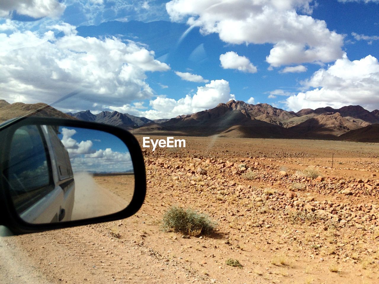 Car reflecting on side-view mirror on road against cloudy sky