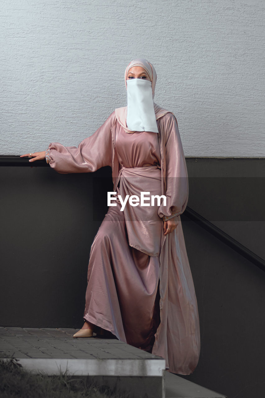 Full length image of veiled woman with white niqab and pink silky dress standing against the wall