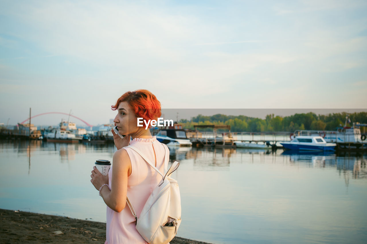 Mid adult woman talking on mobile phone while standing by river against sky during sunset