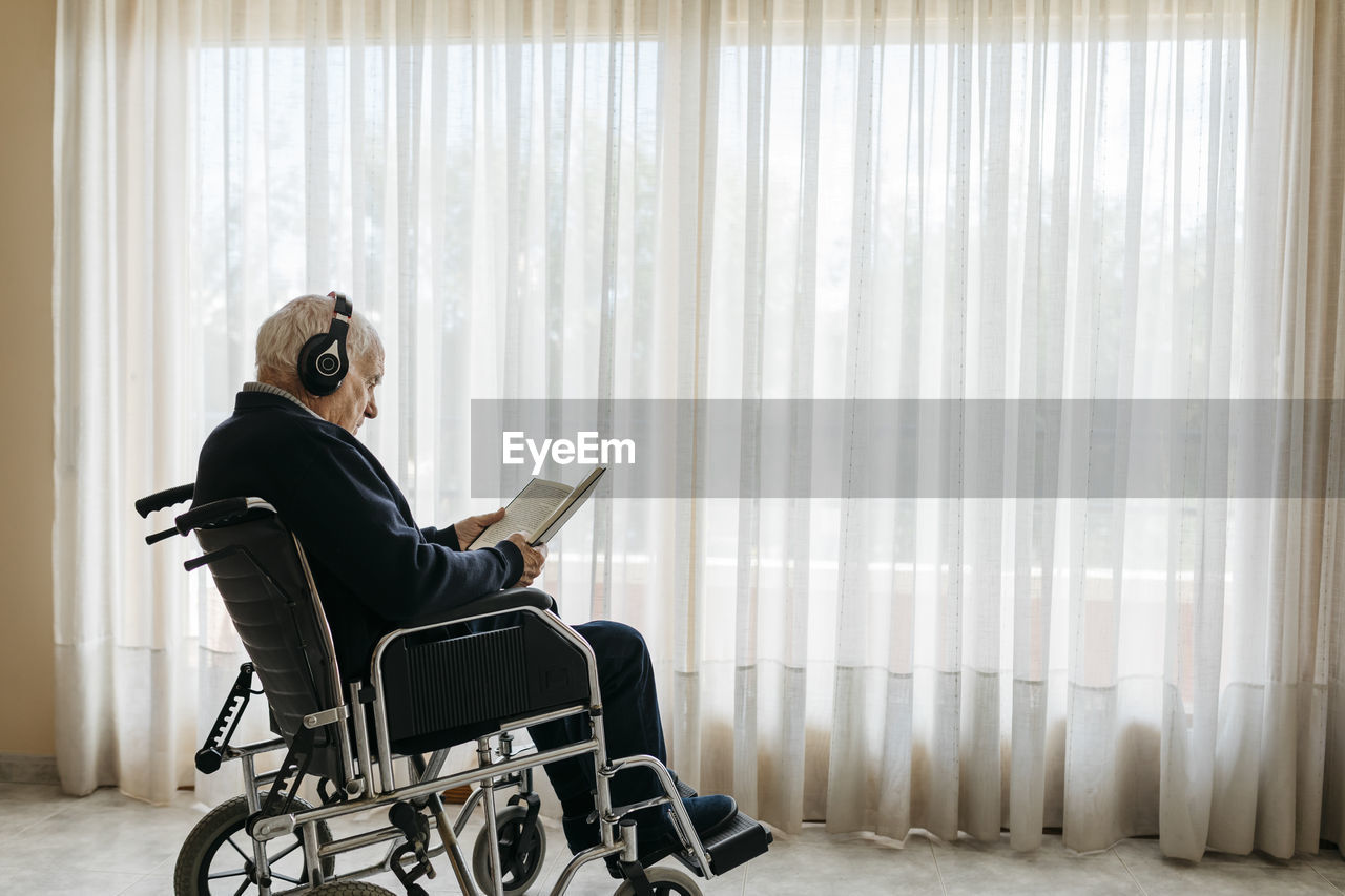 Senior man sitting in wheelchair reading a book while listening music with headphones