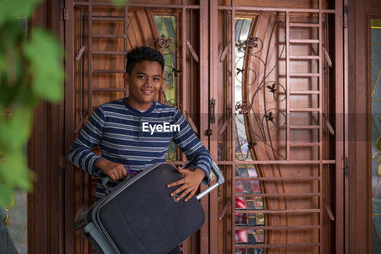 Portrait of smiling boy holding suitcase while standing against door
