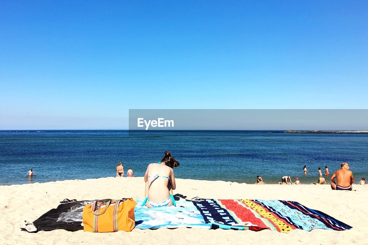 Rear view of woman resting on beach towel at shore against clear blue sky