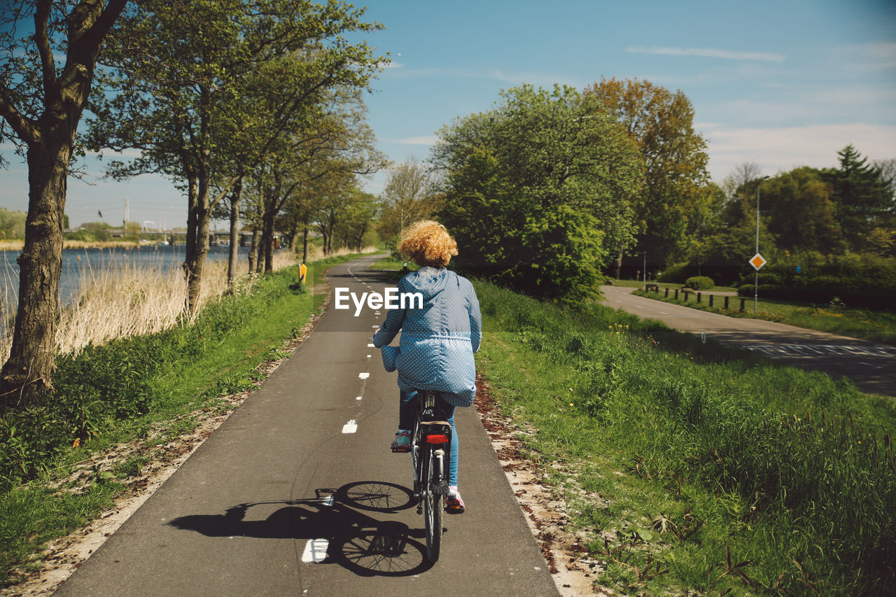 Rear view of woman with curly hair riding bicycle on road during sunny day