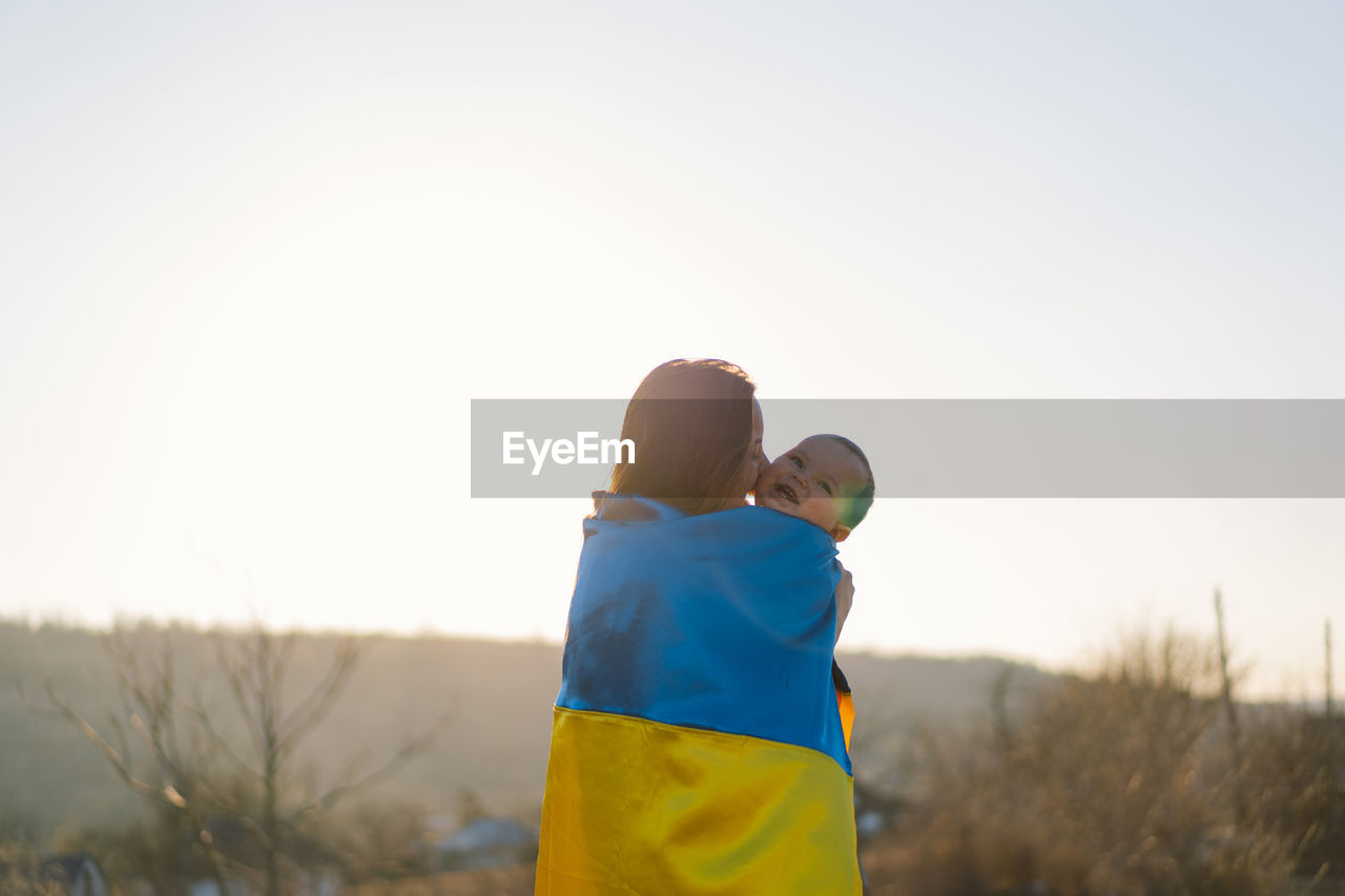 Woman hugs her little son wrapped in yellow and blue flag of ukraine in outdoors.