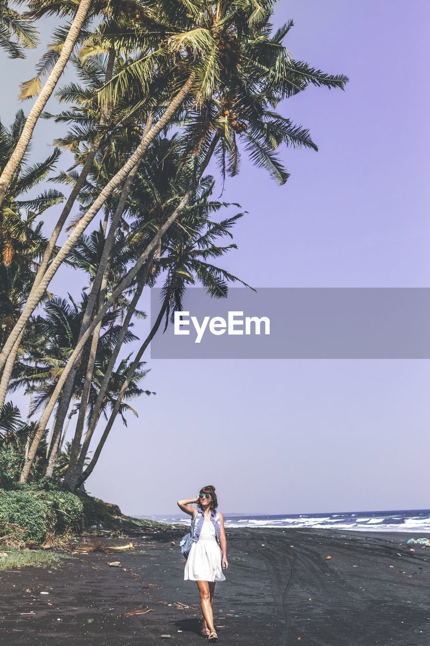 Full length of woman standing by palm tree at beach against sky