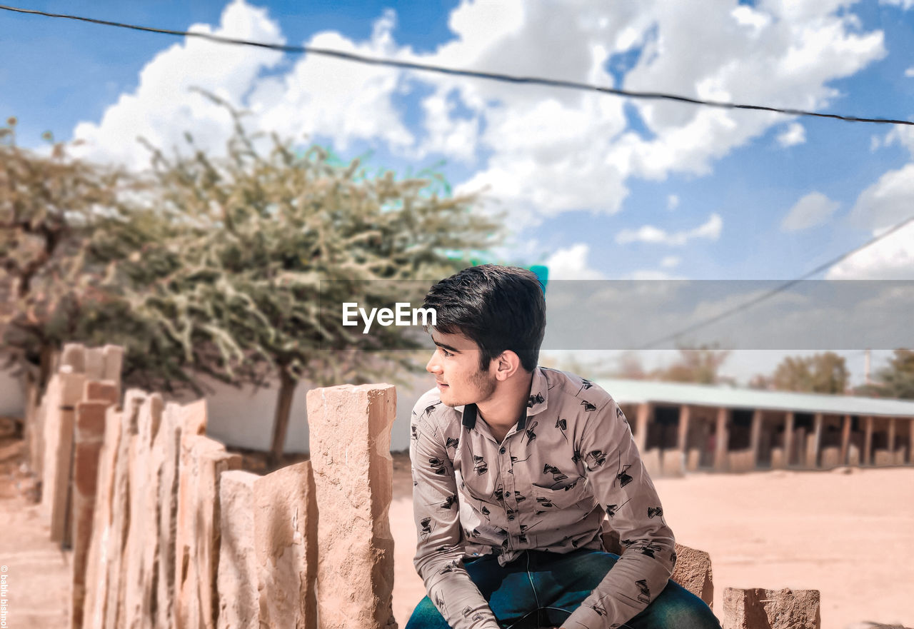 YOUNG MAN LOOKING AWAY WHILE SITTING AGAINST TREES